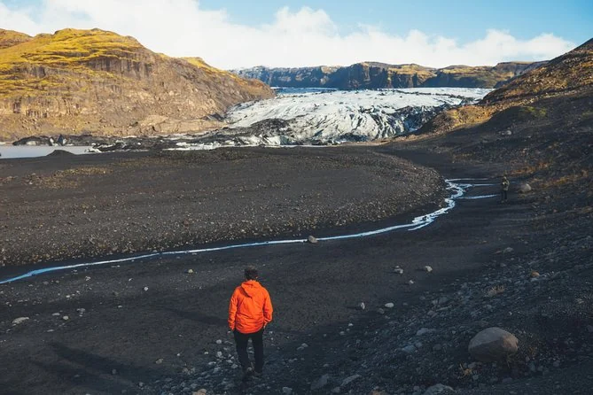 A walker in an orange jacket crossing black gravel and a meltwater stream toward a glacier tongue