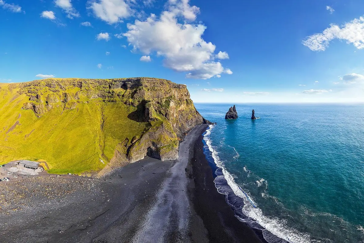 Aerial view of a black sand beach below a green headland, with sea stacks standing offshore