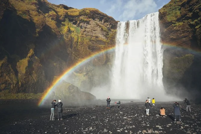 Visitors on black gravel below Skógafoss, with a rainbow arching through the spray