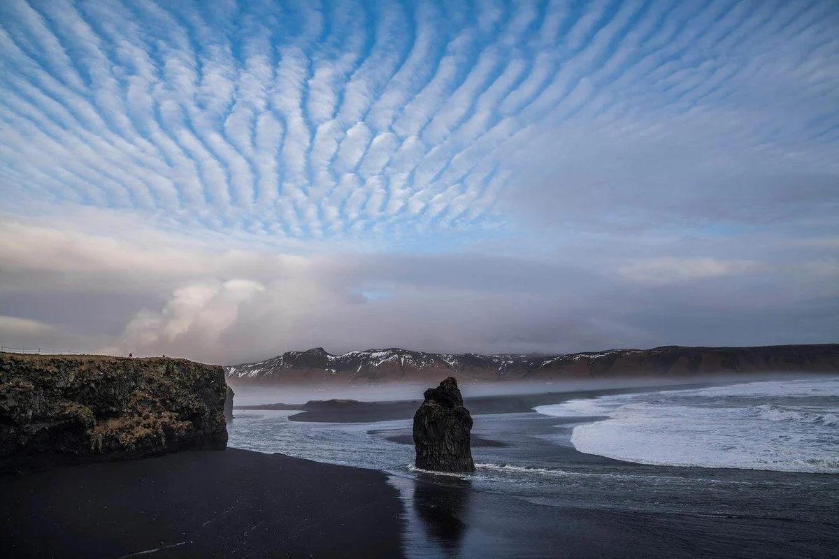 A lone sea stack on a wet black beach under rippled clouds, with a headland on the left