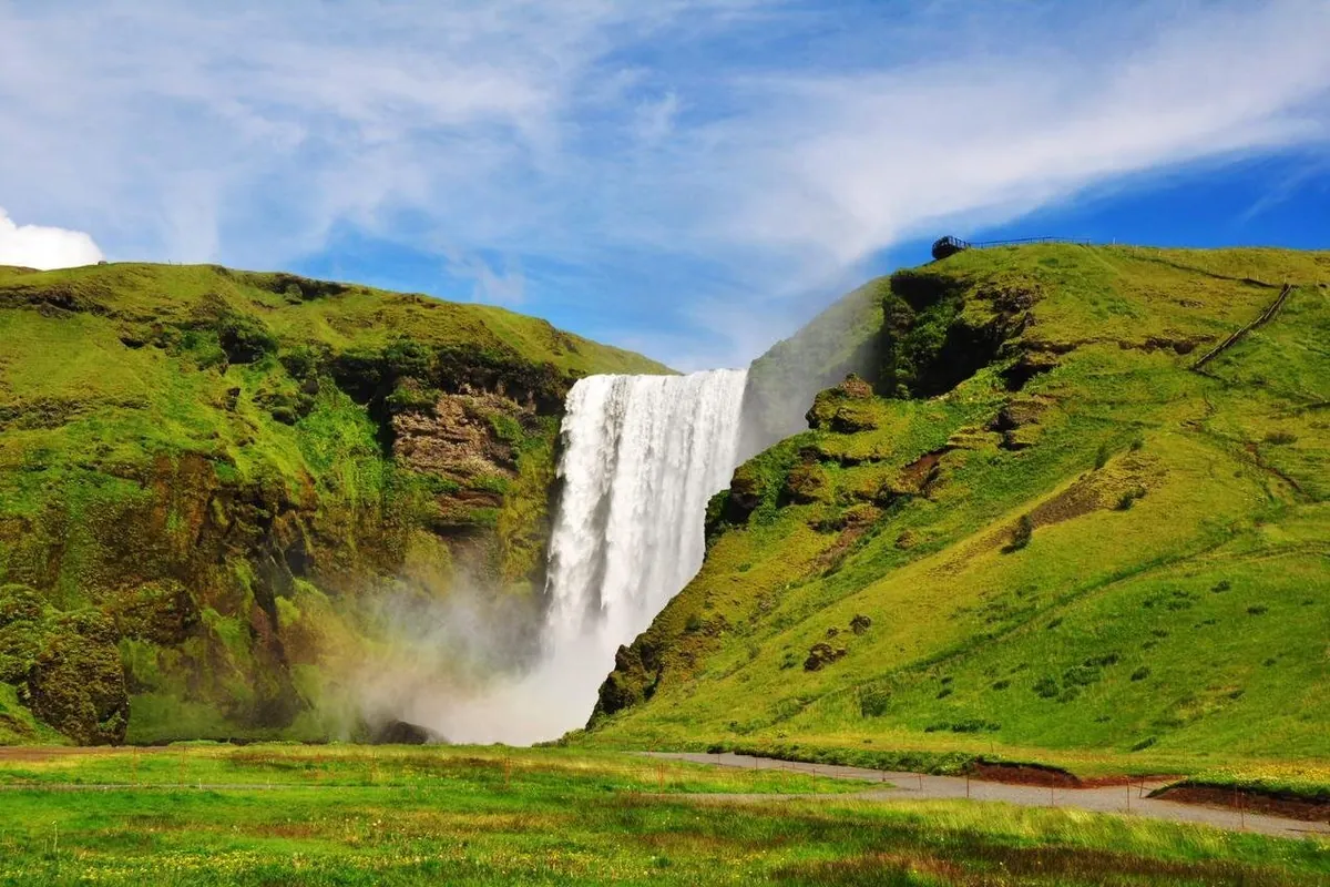 Skógafoss dropping between green slopes, with the staircase path climbing the hill on the right