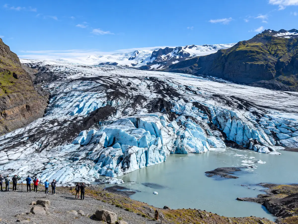 The blue and white tongue of Sólheimajökull with dark ash bands above a meltwater lagoon, and small visitors at a viewpoint