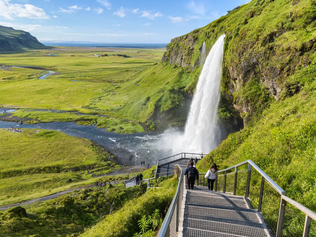 Hikers climbing a metal staircase up a green slope beside a waterfall, with the river and lowland below